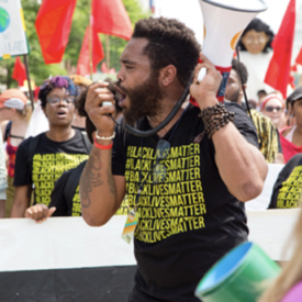 Man with medium skin wearing a Black Lives Matter t shirt and holding a bull horn, speaking at a rally.