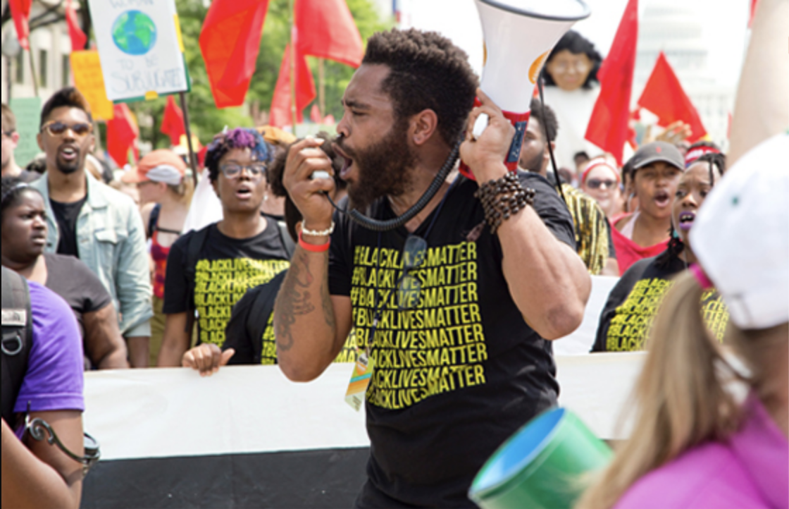 Man with medium skin wearing a Black Lives Matter t shirt and holding a bull horn, speaking at a rally.