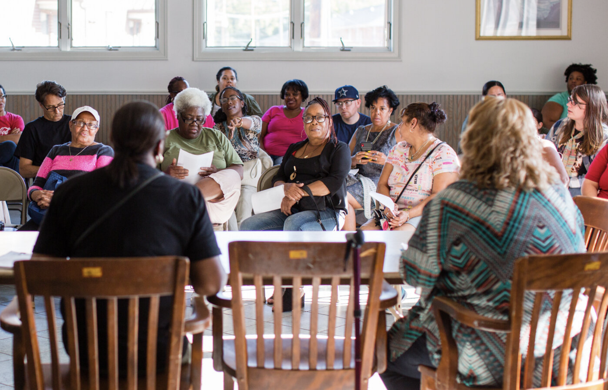 People of various ethnicities and skin tones seated at a meeting in a large, sunny room.