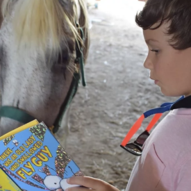 Photo shows a child of about 6 holding a book in front of a white horse whose nose is turned to and up close to the book.