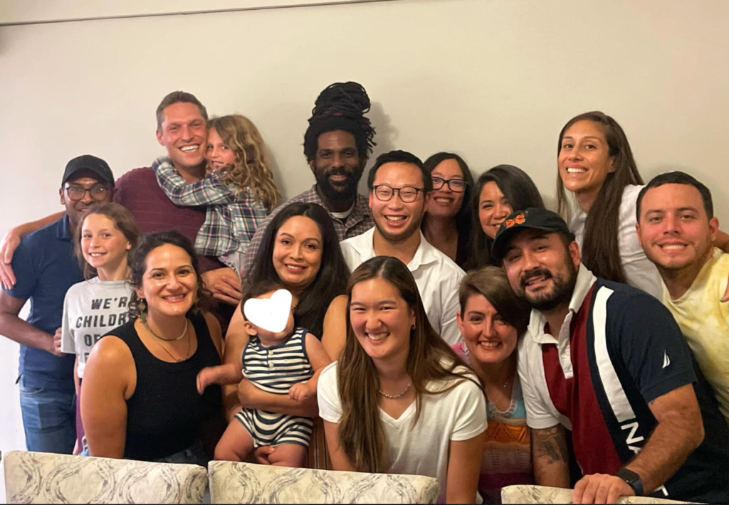 16 people of various ethnicities from children to young adult are shown gathered and smiling for a photo indoors, wearing summer clothing.