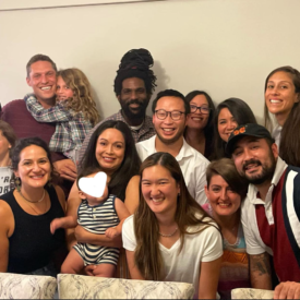 16 people of various ethnicities from children to young adult are shown gathered and smiling for a photo indoors, wearing summer clothing.