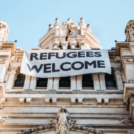 The top part of an old building with ornate carvings is shown, with a banner hanging from it that reads "Refugees Welcome."