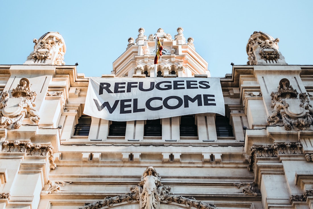 The top part of an old building with ornate carvings is shown, with a banner hanging from it that reads "Refugees Welcome."