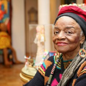 Faith Ringold sitting in the foreground of her art show. She is an elderly black woman with grey locs