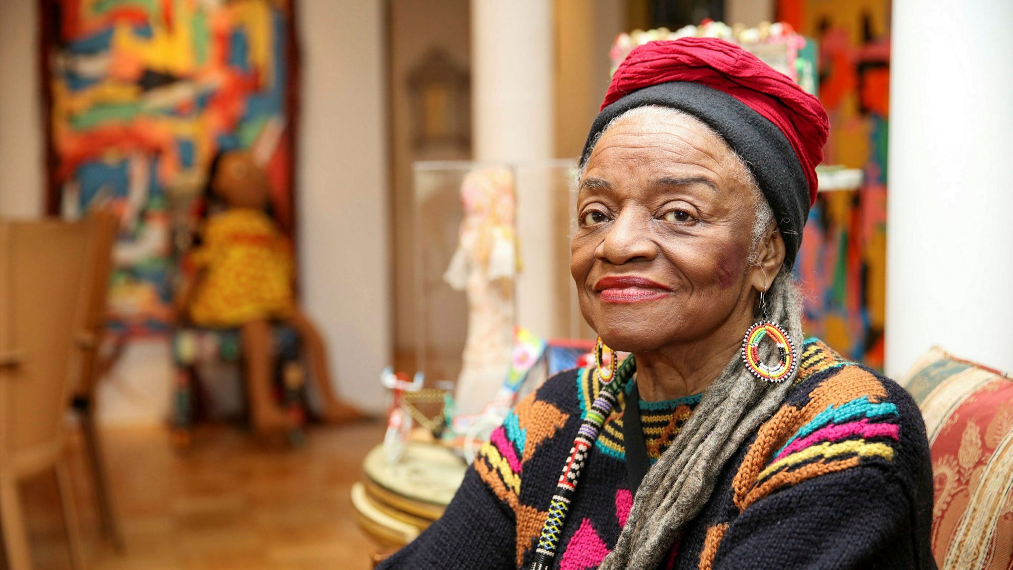 Faith Ringold sitting in the foreground of her art show. She is an elderly black woman with grey locs