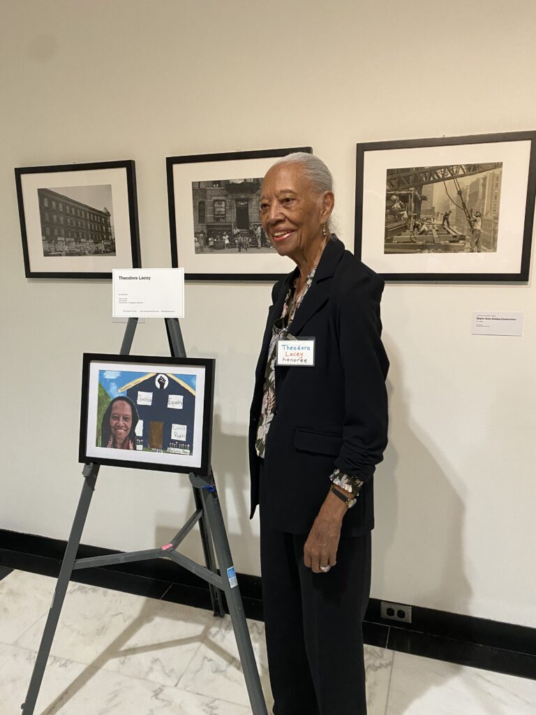 Theodora Lacey is an elderly Black woman with grey hair in a pony tail wearing a black suit smiling with an artistic photo of herself.