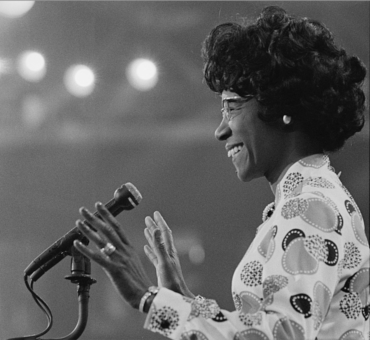 Photograph shows Shirley Chisholm smiling at microphone.