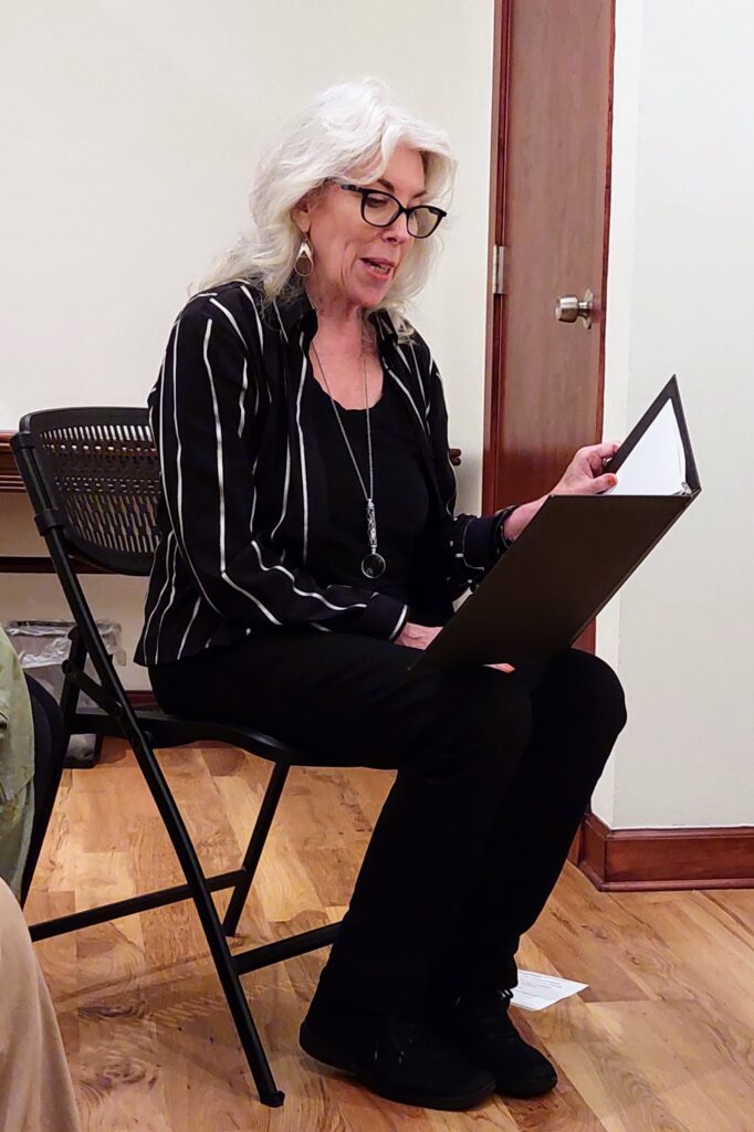 Seated woman with grey hair and glasses reading aloud