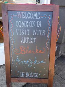 a side walk sign saying Welcome Come On In Visit With Artist Blanka Amezkua