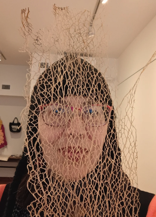 Photo of a woman wearing a close fitting hat and glasses, seen behind the lacy structure of a dried nopal plant
