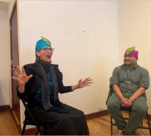 Blanka and Marco in front of the room wearing nopales on their foreheads held on with blue and magenta headwraps